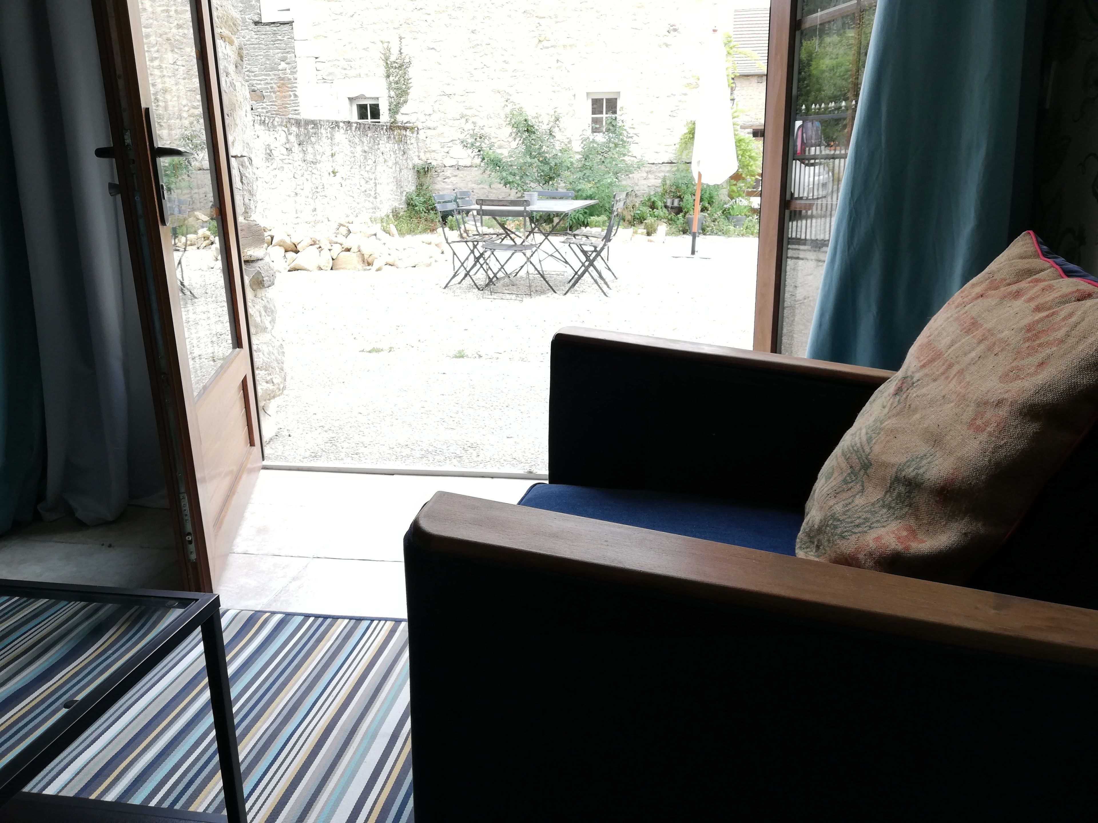 A room with a sofa and a table, a view of an outdoor area with a table and chairs, and a stone building in the background.