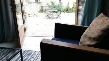 A room with a sofa and a table, a view of an outdoor area with a table and chairs, and a stone building in the background.