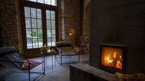A cozy room with a stone fireplace, a comfortable chair, a small table with a candle, and a view through large windows. - Au Domaine de la Pommeraye & Spa (La Pommeraye)