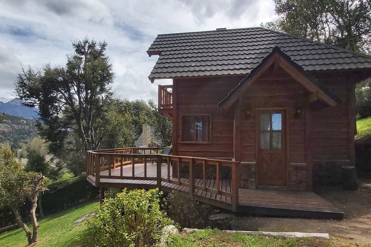Une cabane en bois dotée d’un balcon, entourée d’arbres et offrant une vue sur un paysage montagneux.