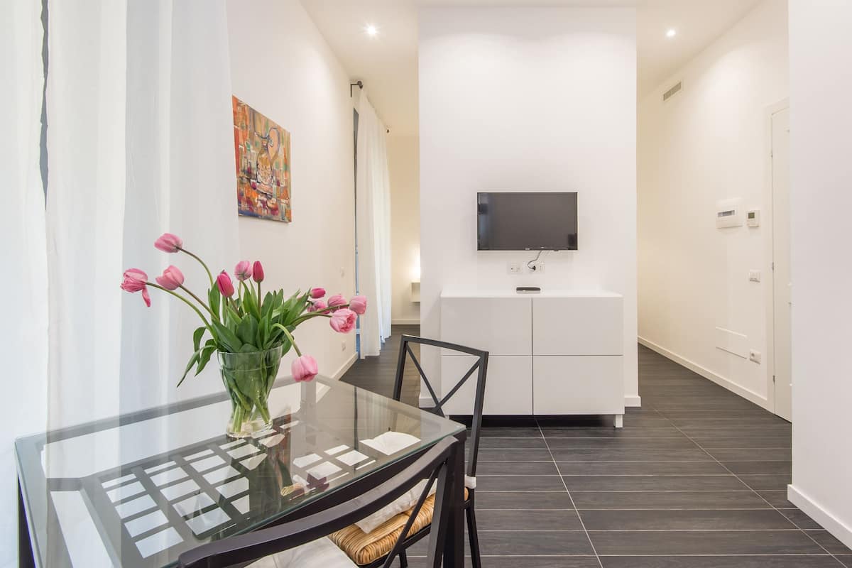A modern dining area with a glass table, chairs, and a vase of pink tulips. A flat-screen TV is mounted on the wall above a white cabinet.