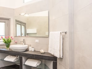 A modern bathroom with a large mirror, white sink, and dark countertop. There are two towels on a glass shelf and a vase with flowers on the counter. - Residenza Ottaviano 105 by Hili (Rome)