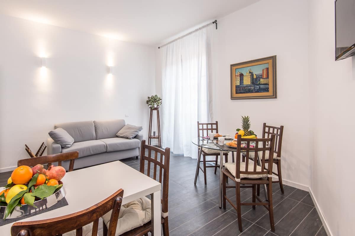 A modern living room with a grey sofa, a dining table with chairs, and a painting on the wall.
