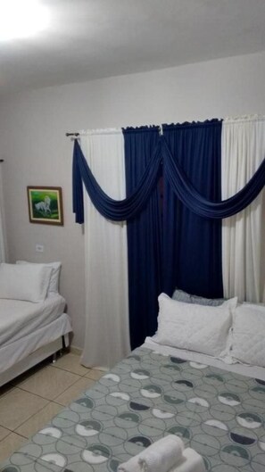A hotel room with two beds, one with a patterned bedspread and the other with plain bedding. There is a curtain divider with blue and white drapes. A framed picture hangs on the wall. - Pousada Palace Nook (Olímpia)