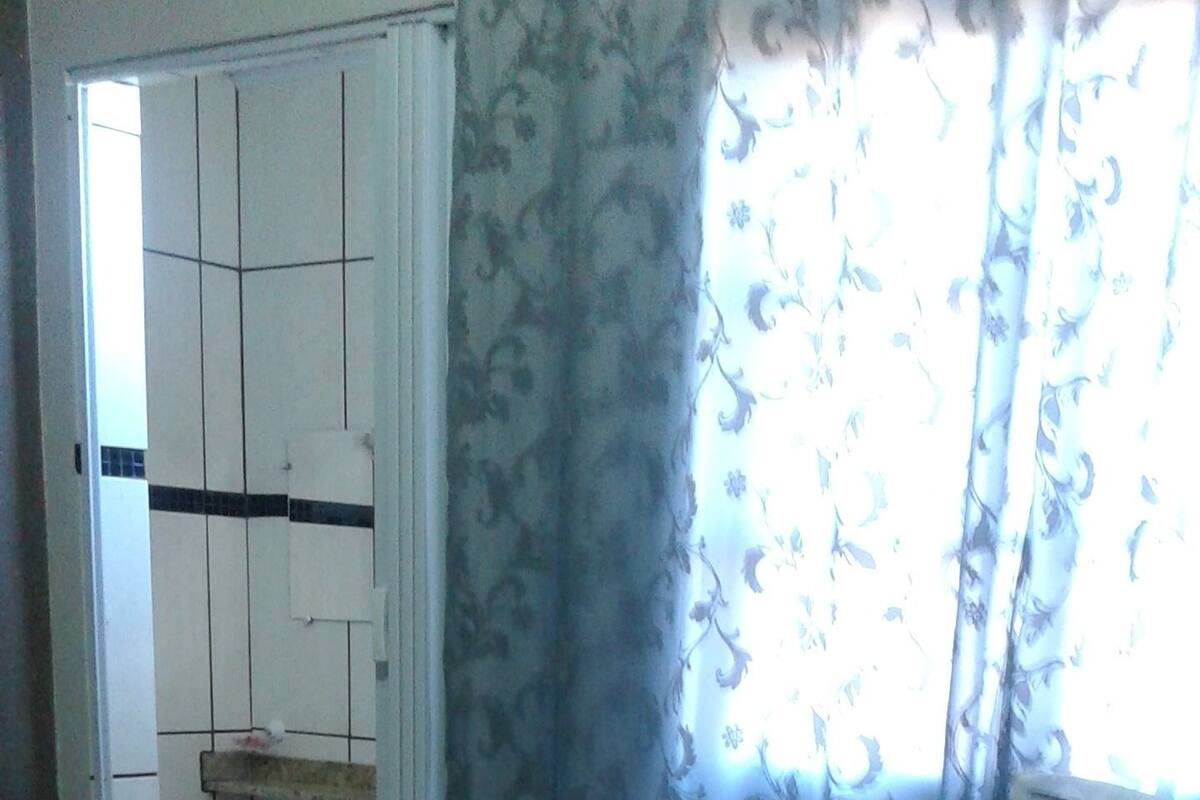 Ein Schlafzimmer mit einem Bett, einem kleinen Tisch mit Gegenständen, einem Kleiderschrank und einem Fenster mit Vorhängen.