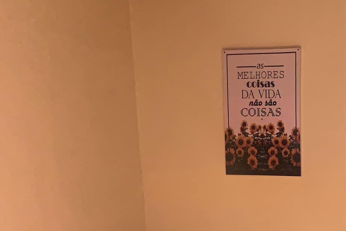 Ein kleines Zimmer mit einem Einzelbett, einer bunten Bettdecke, Holzboden und einem an der Wand befestigten Schild mit Text.