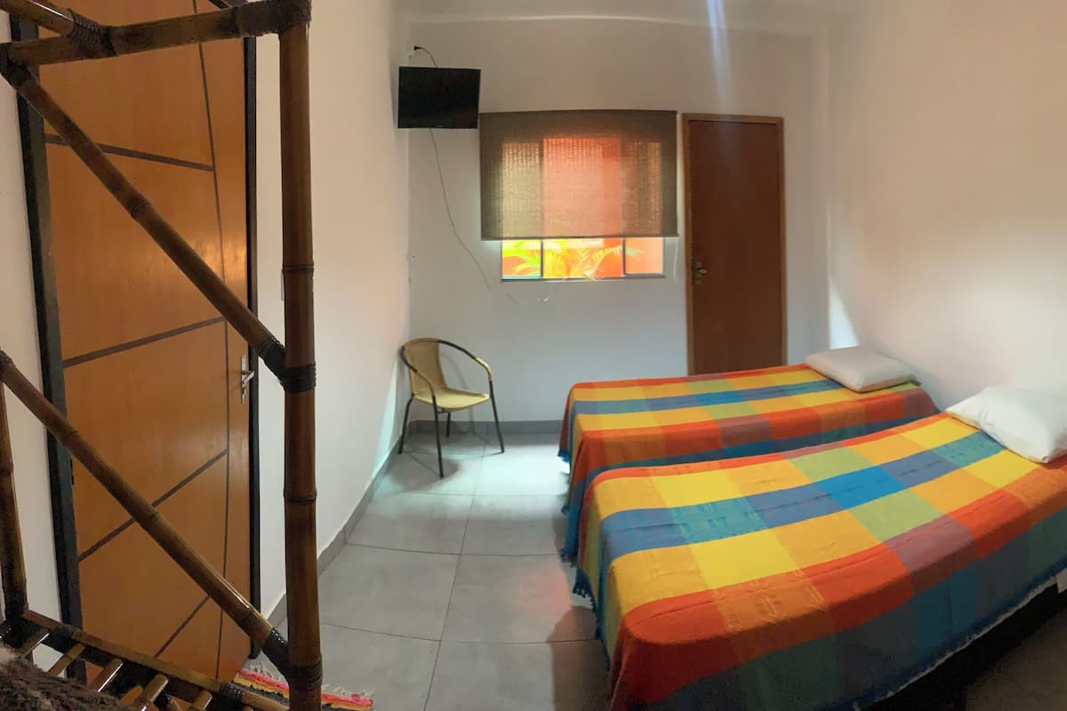 Ein Zimmer mit zwei Betten, einem Sessel, einem Fenster mit Vorhängen und einem an der Wand befestigten Fernseher.