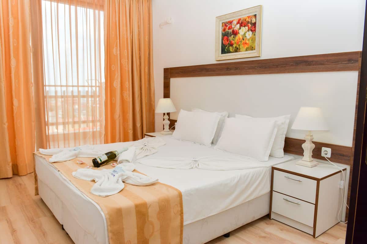 A hotel room with a bed, two bedside tables, a nightstand, a window with curtains, and a painting on the wall.