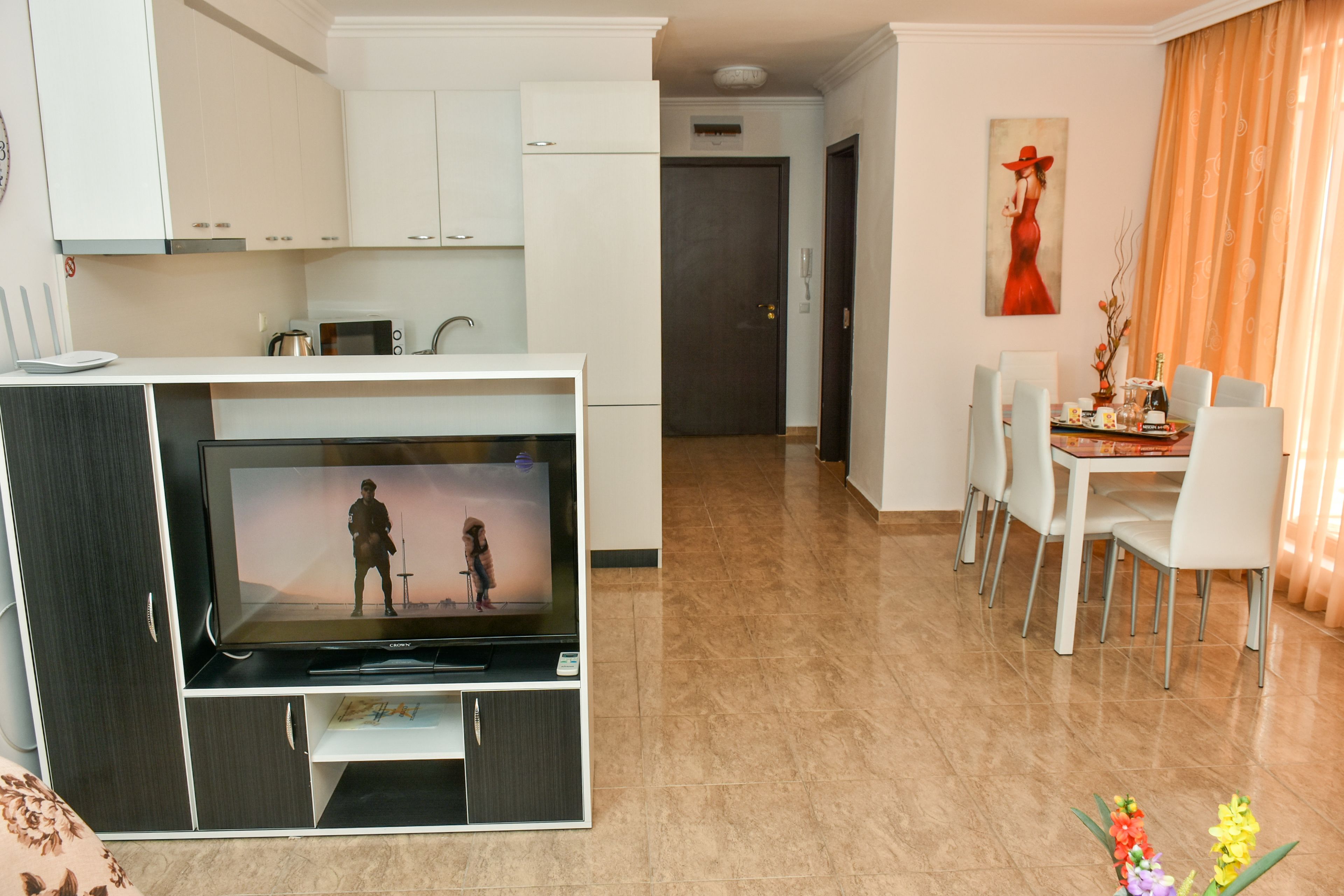 A modern living room with a flat-screen TV, a dining area with white chairs, and a kitchen with white cabinets.