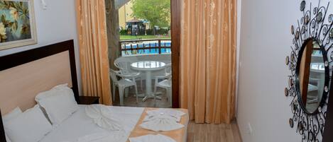 A hotel room with a bed, a small table, a chair, and a view of an outdoor area with a pool and umbrellas.