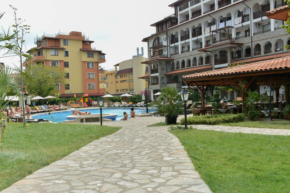 A pool area with lounge chairs and umbrellas, surrounded by buildings with balconies and a stone pathway.