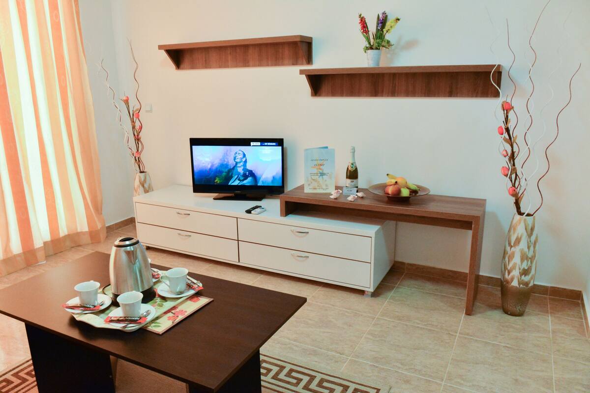 A modern living room with a television, a coffee table with cups and a book, a wooden desk with a plant, and a decorative vase.