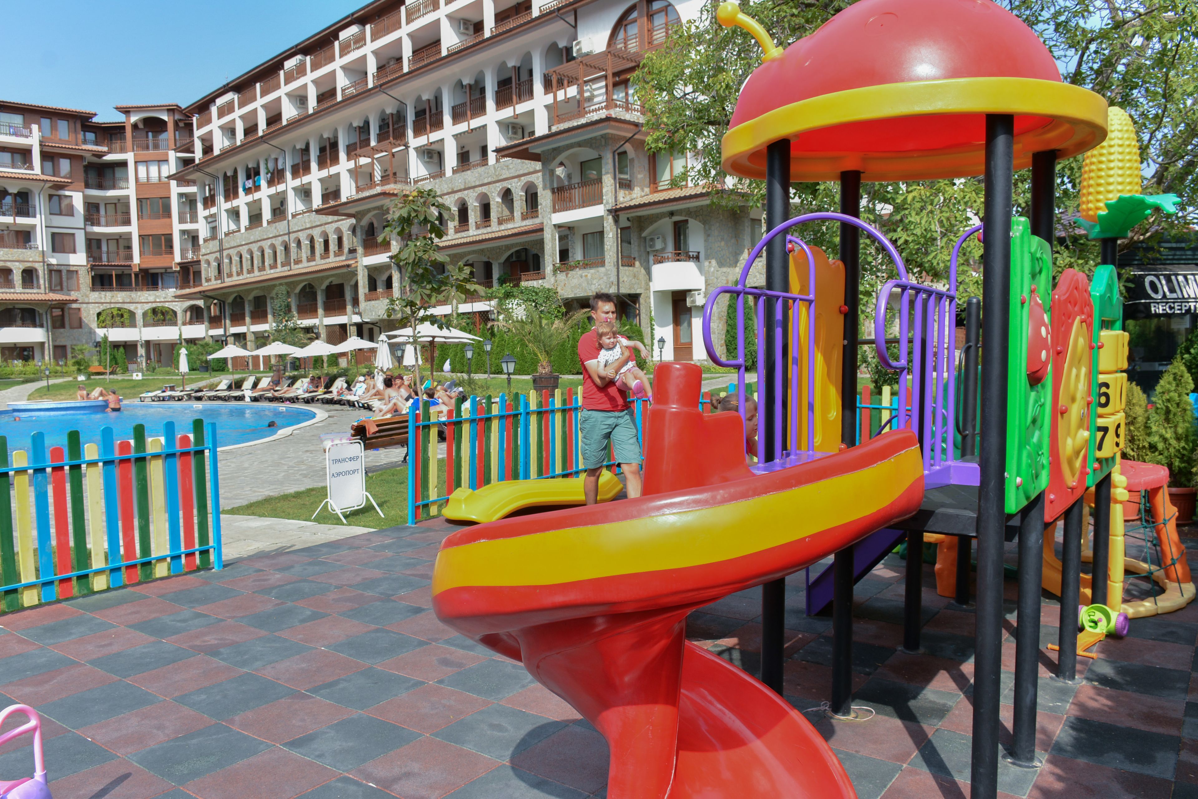 A playground with a slide and climbing structures, surrounded by a pool and a hotel with multiple balconies.