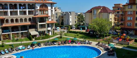 A swimming pool surrounded by lounge chairs and umbrellas, with multi-story buildings in the background.