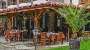 Un área de comedor al aire libre con suelo de piedra, mesas y sillas de madera, y una zona de estar cubierta.