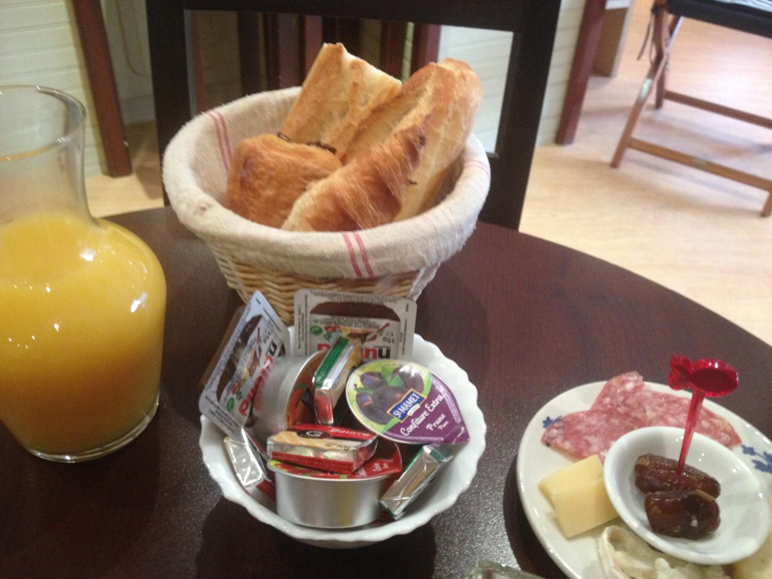 Una escena de desayuno con una cesta de croissants, un vaso de zumo de naranja y un plato con queso, jamón y una pequeña fuente de mermelada.