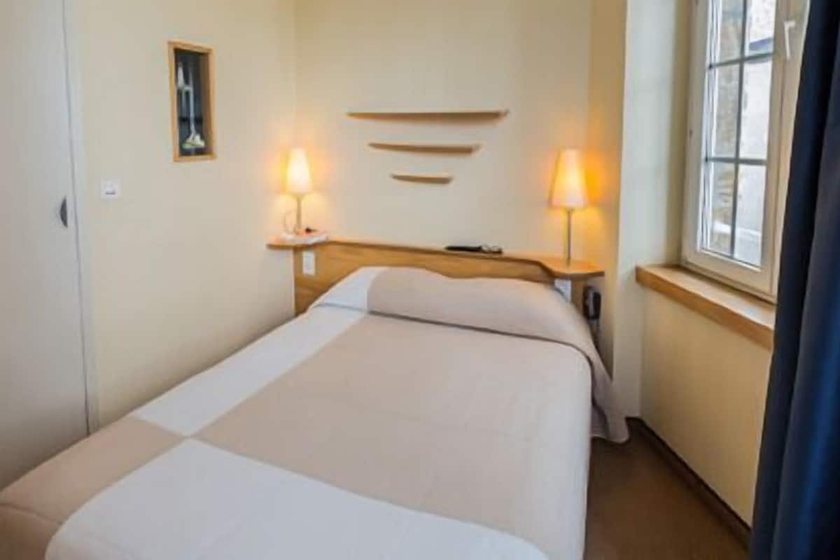 Una habitación de hotel con cama, lámparas de noche, una ventana con cortinas y una repisa en la pared.