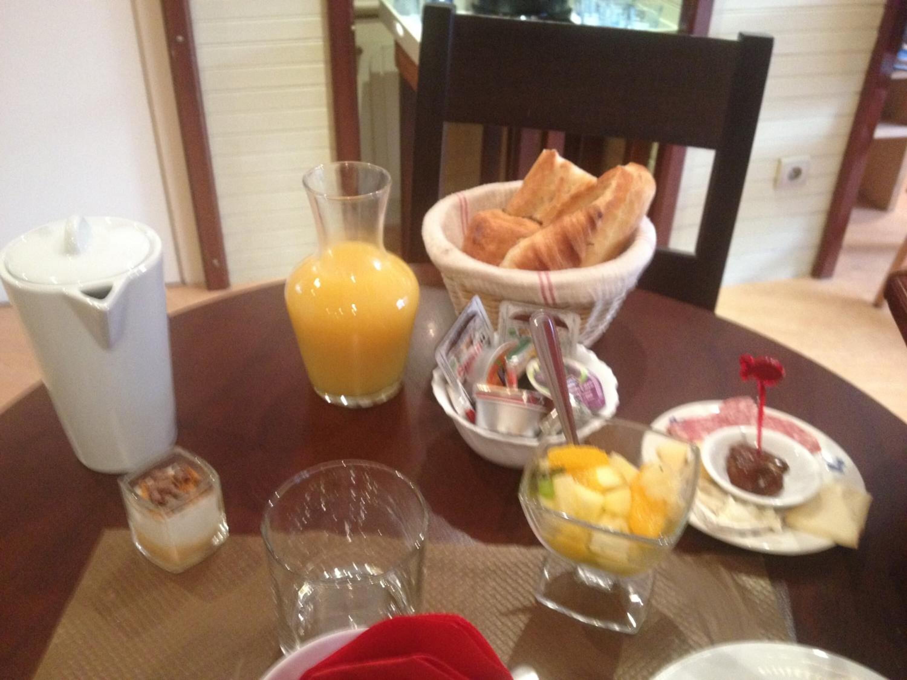 A breakfast table with a pitcher of orange juice, a bowl of fruit, a basket of croissants, a small glass of milk, and a plate with jam and toast.