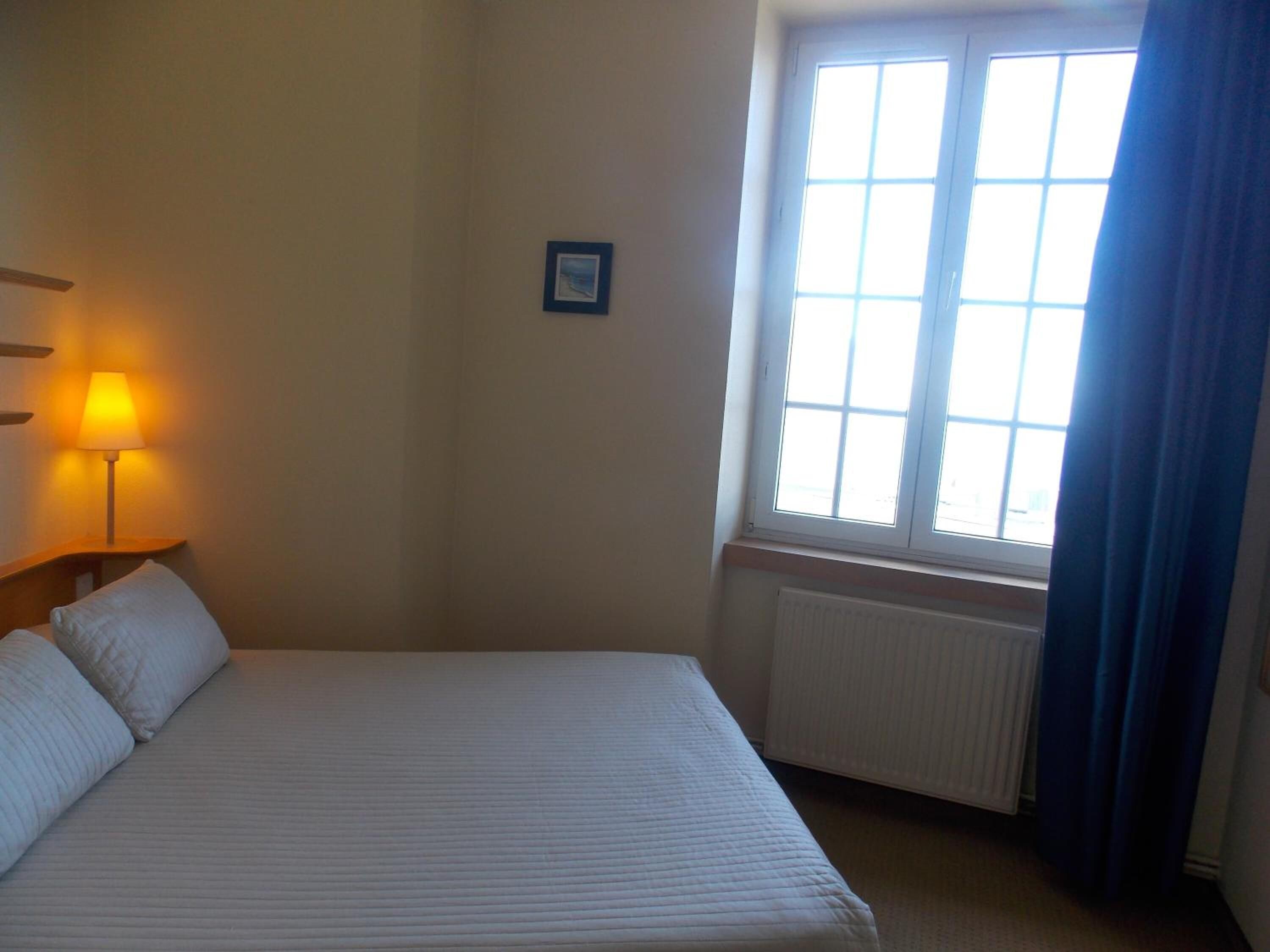 Une chambre d’hôtel avec un grand lit, une fenêtre avec des rideaux, un radiateur et un tableau accroché au mur.