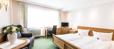 A hotel room with a large bed, a desk with a computer, a chair, a sofa, and a vase of flowers.
