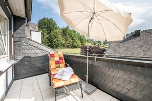 A balcony with a chair, a large umbrella, and a view of a garden and buildings. - Landgasthof Hotel Hanses-Bräutigam (Schmallenberg)