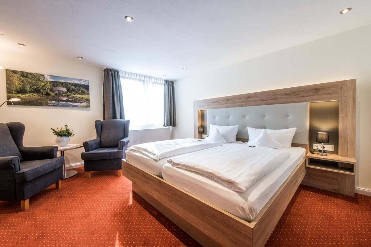 Une chambre d’hôtel avec un grand lit, deux fauteuils, une table de chevet, une lampe et un tableau au mur.