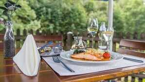A wooden table with a white napkin, a fork, a glass pitcher, a glass of white wine, and a plate with salmon and vegetables. - Landgasthof Hotel Hanses-Bräutigam (Schmallenberg)