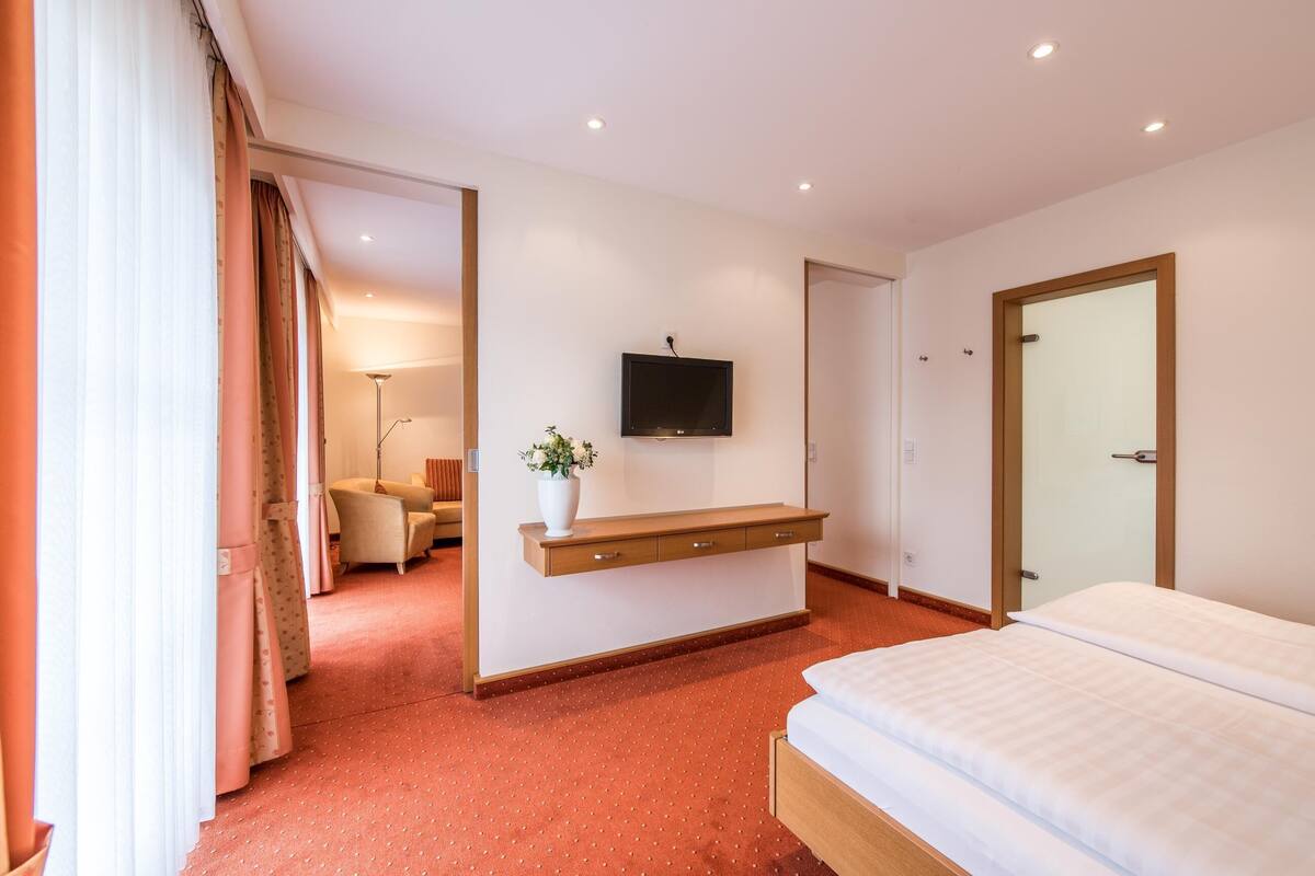 Une chambre d’hôtel avec un lit, un téléviseur fixé au mur, une table de chevet ornée d’un vase de fleurs et une chaise dans un coin.