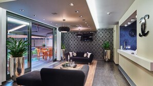 A modern living room with a black sofa, a coffee table, and a dining area with chairs and a table. There is a large window with plants and a view of the outside. - Boutique Hotel Chrysso (Kosice)