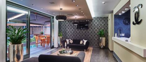 Una sala moderna con un sofá negro, una mesa de centro y un comedor con sillas y una mesa. Hay un ventanal con plantas y vistas al exterior.