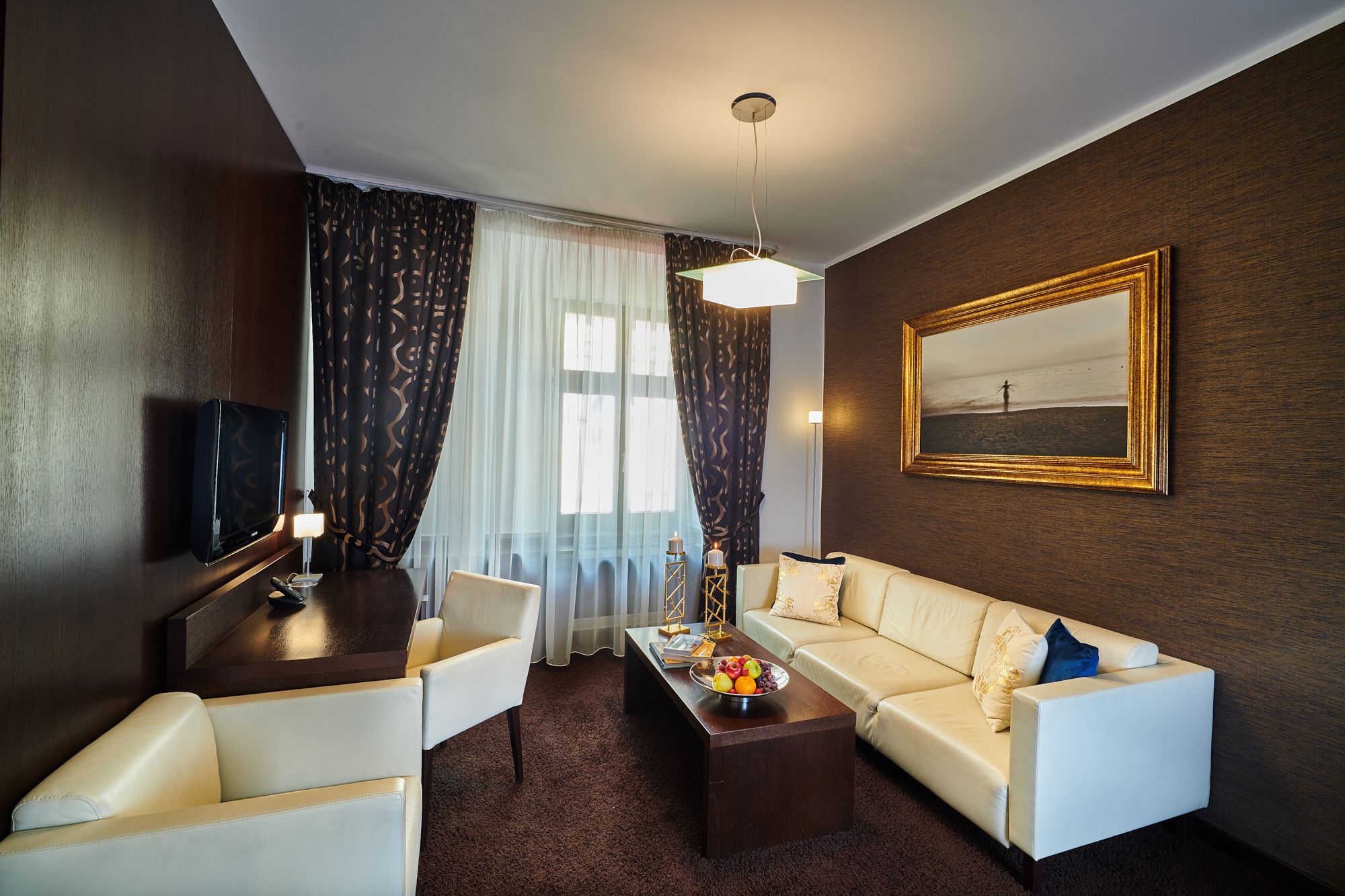 Una habitación de hotel con sofá, sillones, una mesa de centro con fruta, televisor, un cuadro y un amplio ventanal con cortinas.