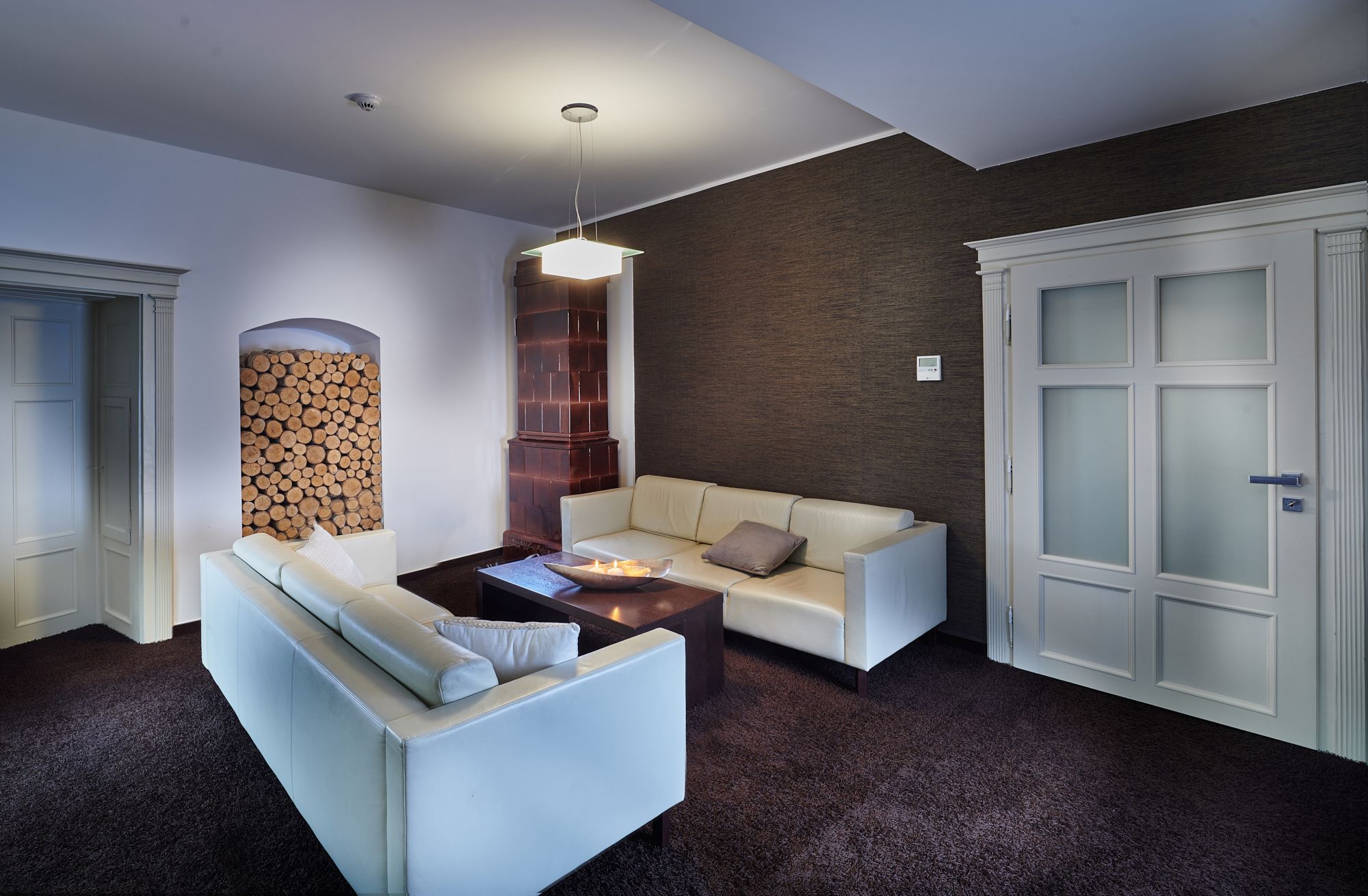 A modern living room with a white sofa, a coffee table, and a fireplace. There is a stack of firewood against the wall and a door with frosted glass panels.