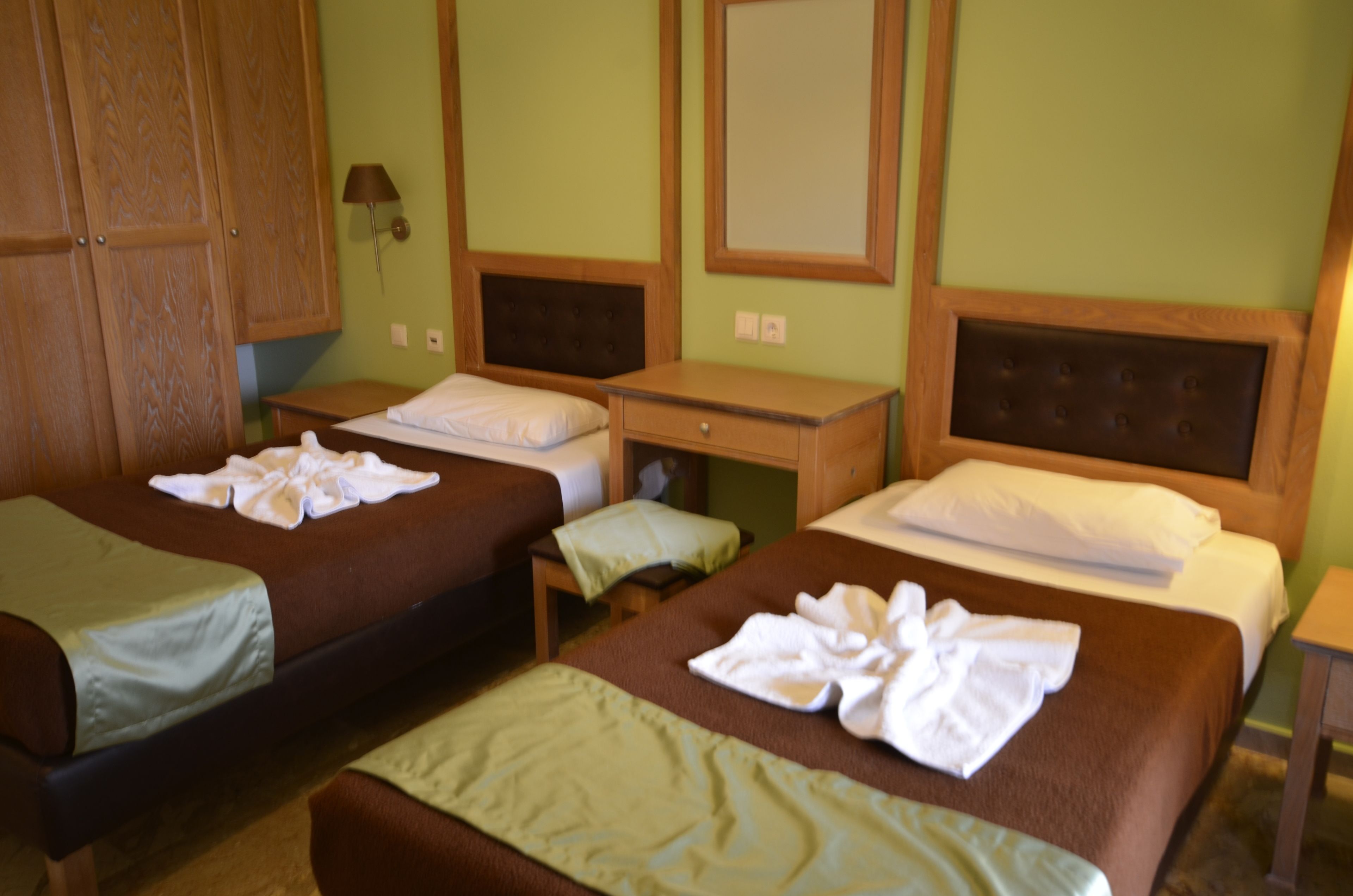 A hotel room with two single beds, a wooden wardrobe, a bedside table with a lamp, and a mirror on the wall.