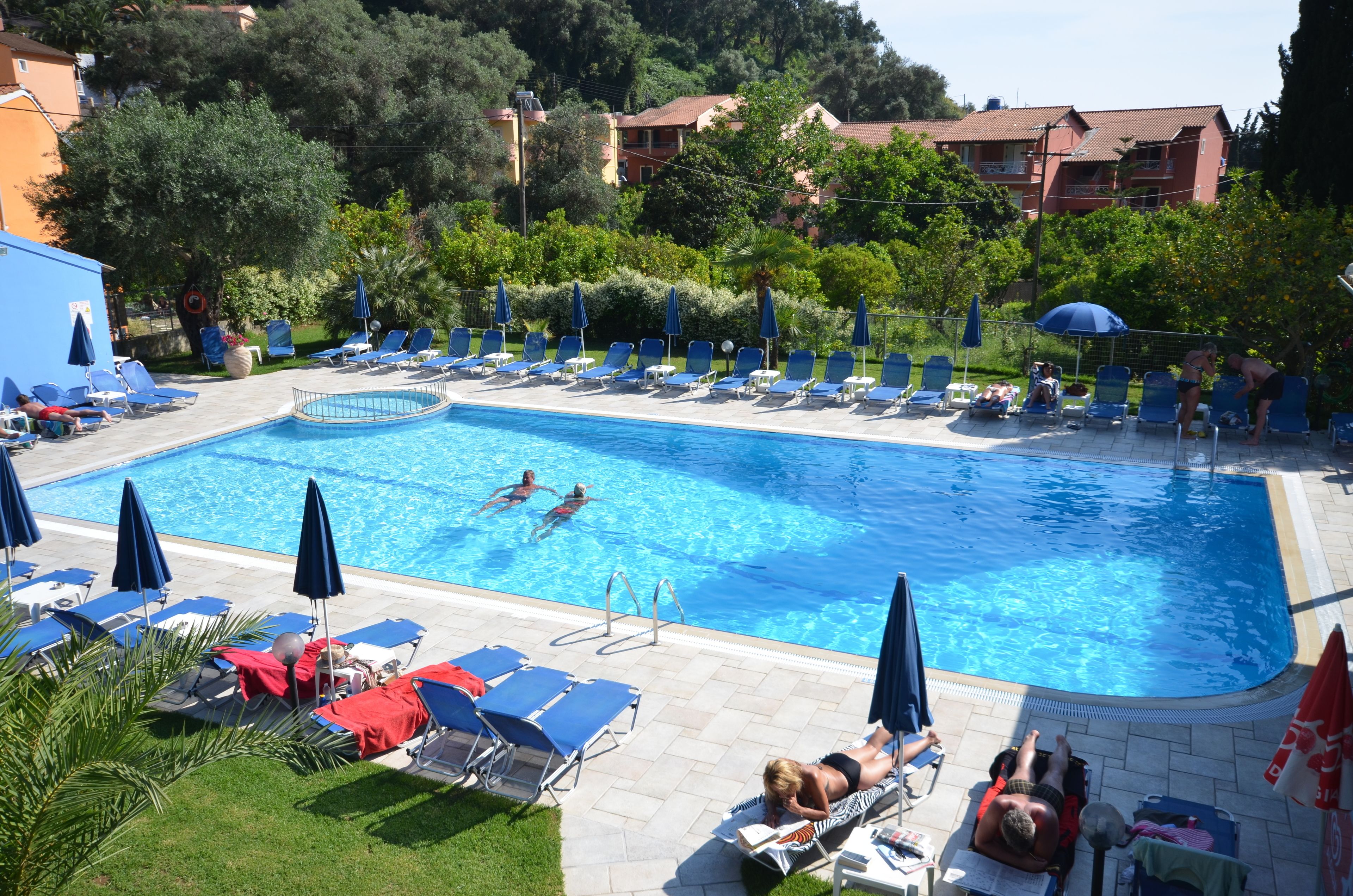 A swimming pool with lounge chairs and umbrellas, surrounded by greenery and buildings.