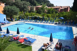 Una piscina con sillas de descanso y sombrillas, rodeada de follaje y edificios.