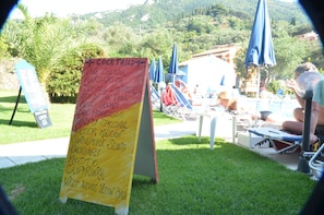 A cocktail menu board outdoors with a pool area in the background. - Irene Apartments (Corfu)
