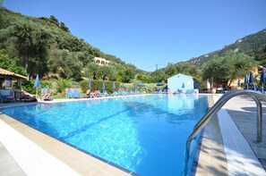 A swimming pool with lounge chairs and umbrellas, surrounded by greenery and hills. - Irene Apartments (Corfu)