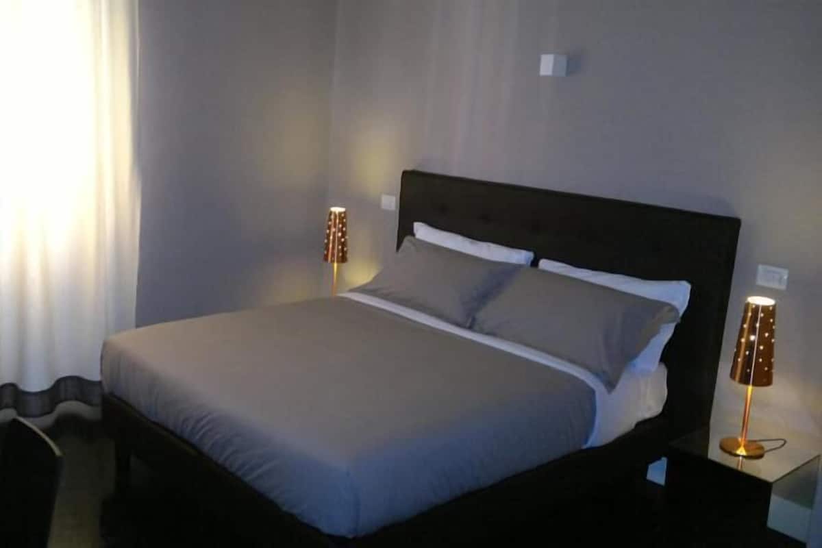 A neatly made bed with a grey duvet, two bedside lamps, and a window with curtains.