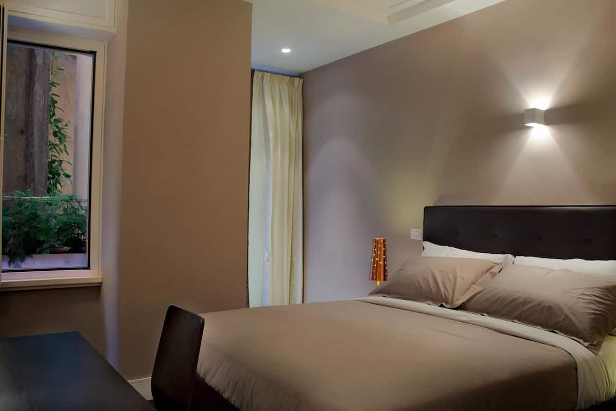 A bedroom with a bed, a window with a view of greenery, a table, and a wall-mounted light fixture.