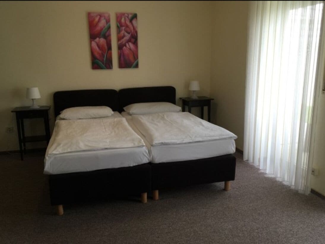 Cama doble con estructura negra y ropa de cama blanca en una habitación con paredes color beige. Hay dos mesitas de noche con lámparas y una ventana con cortinas traslúcidas.