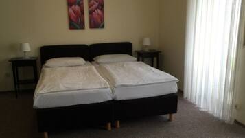 Cama doble con estructura negra y ropa de cama blanca en una habitación con paredes color beige. Hay dos mesitas de noche con lámparas y una ventana con cortinas traslúcidas.