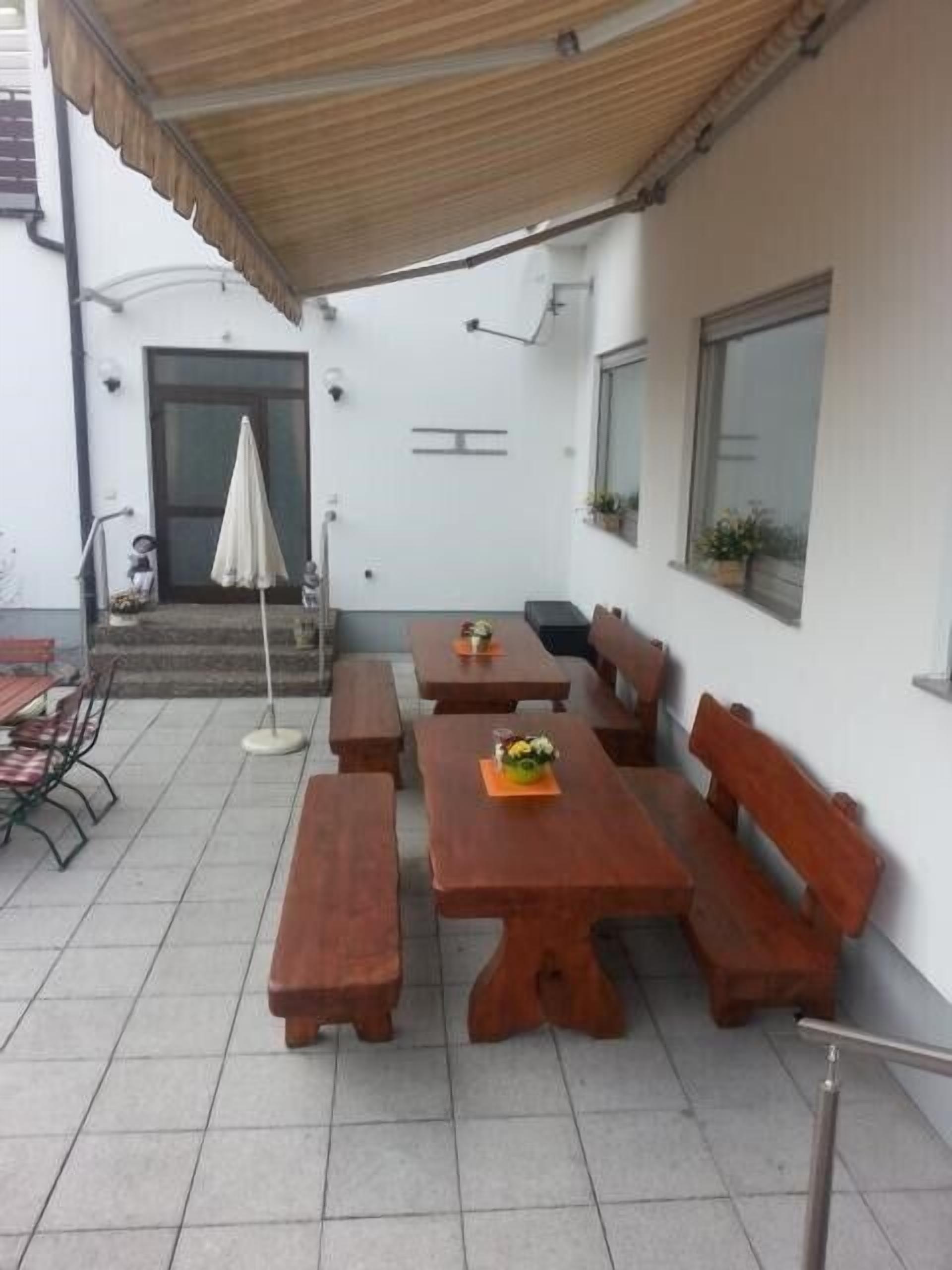 Un espace extérieur aménagé en terrasse, avec des tables et des bancs en bois, un coin salon abrité et une porte donnant accès à l’intérieur.