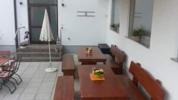 Un espace extérieur aménagé en terrasse, avec des tables et des bancs en bois, un coin salon abrité et une porte donnant accès à l’intérieur.