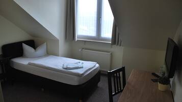 Une petite chambre avec un lit simple, un plafond en pente, une fenêtre avec des rideaux, un radiateur, un bureau en bois et une chaise.