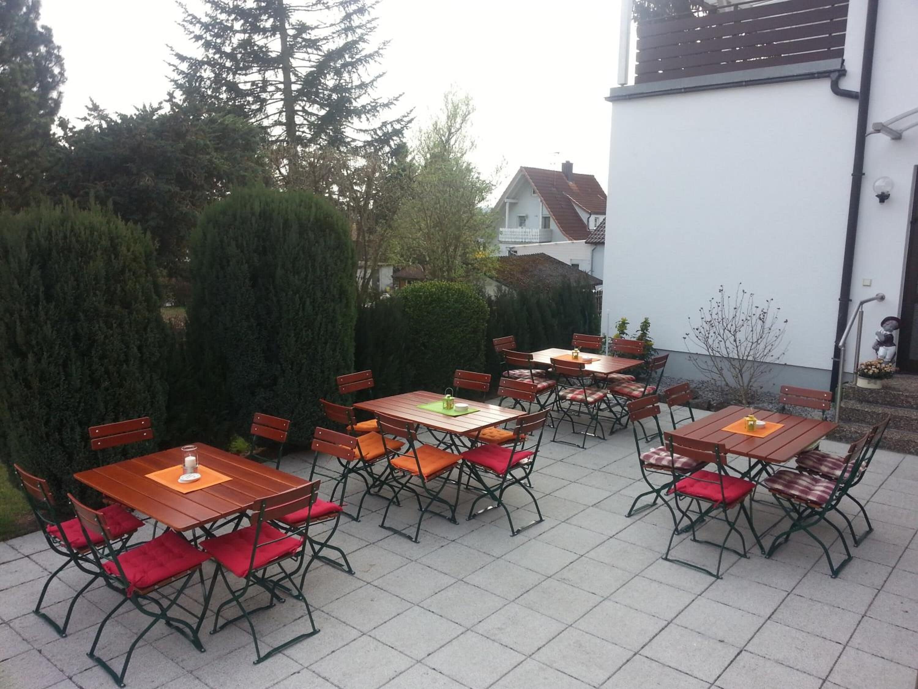 An outdoor patio with multiple wooden tables and chairs, surrounded by greenery and a white building.