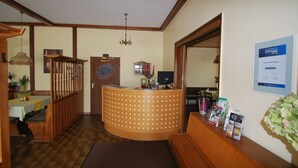 Zona de recepción con un mostrador de madera, una araña, un mostrador de registro y un tablón de anuncios con un cartel promocional de temporada.