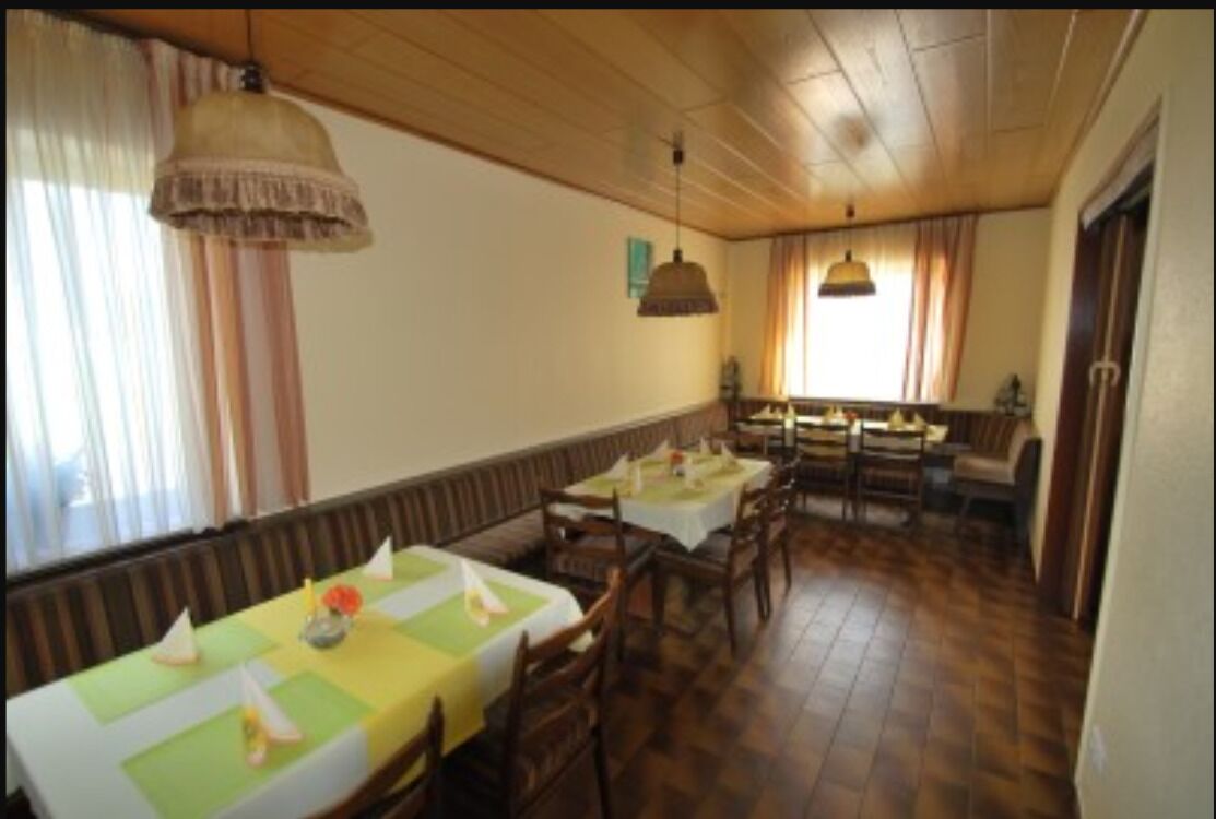 A dining area with wooden tables and chairs, a high ceiling, and a window with curtains.