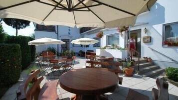 An outdoor patio area with round wooden tables and chairs, covered by large umbrellas.