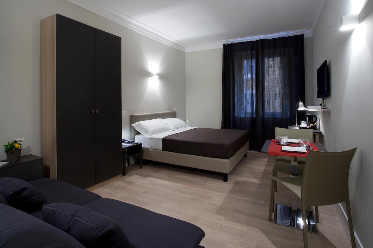 A hotel room with a bed, a desk with a chair, a wardrobe, a TV, and a window with curtains.
