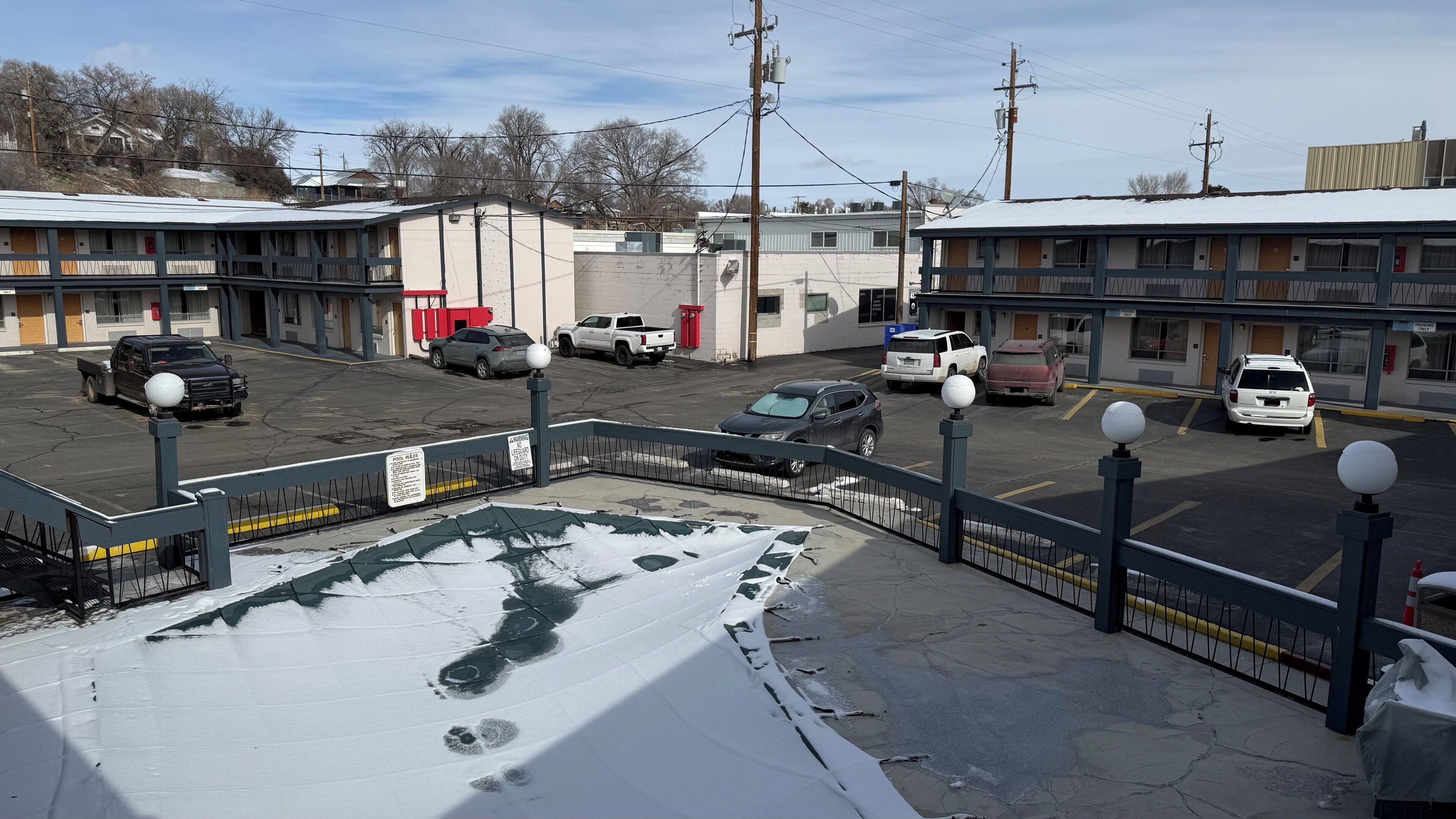 A parking lot with several cars, a building with a sign, and a fenced area with snow.